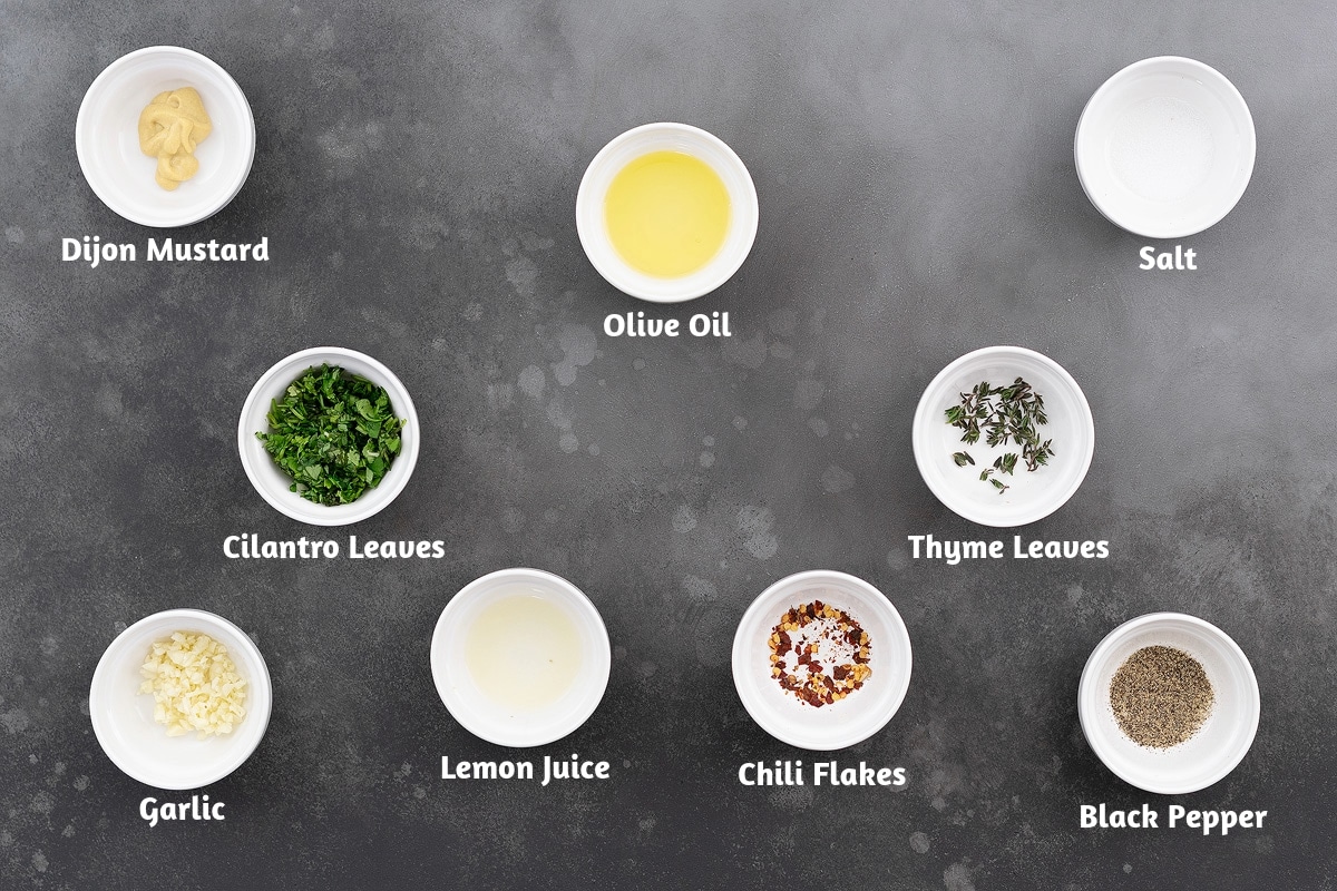 Ingredients for lemon dressing including Dijon mustard, olive oil, salt, cilantro leaves, thyme leaves, garlic, lemon juice, chili flakes, and black pepper powder on a kitchen surface.