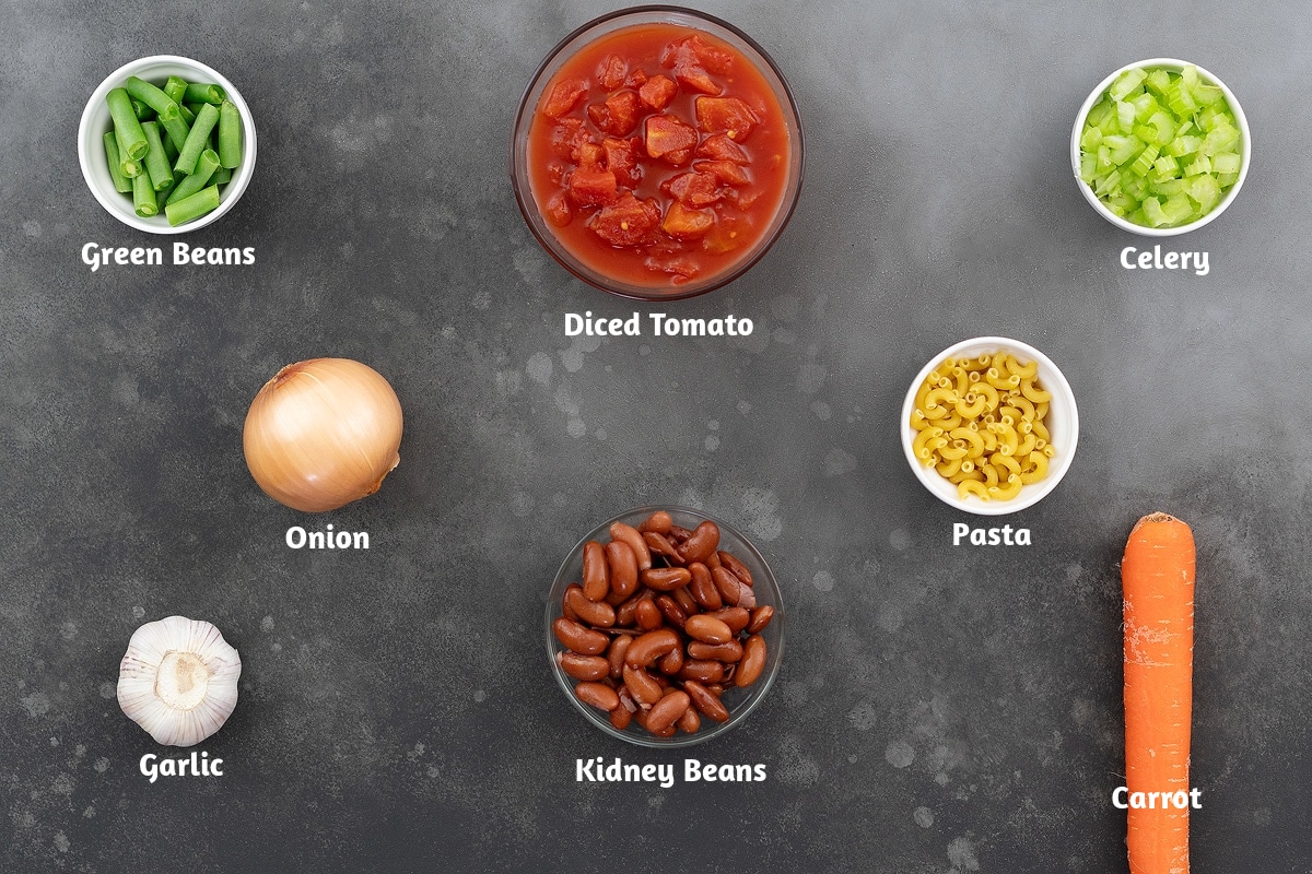 Ingredients for minestrone soup neatly arranged on a gray table, including green beans, diced tomato, celery, onion, pasta, garlic, kidney beans, and carrot.