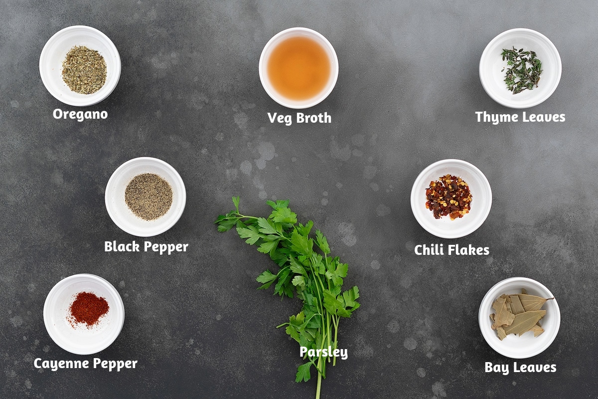 Seasonings and herbs for minestrone soup, including oregano, vegetable broth, thyme leaves, black pepper powder, chili flakes, cayenne pepper, parsley, and bay leaves, displayed on a gray table.