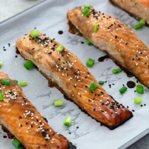 Miso-glazed salmon on a baking sheet garnished with black and white sesame seeds and spring onions.