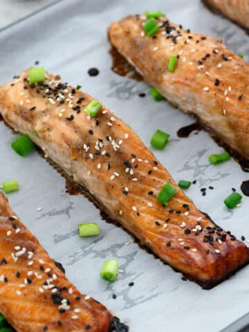 Miso-glazed salmon on a baking sheet garnished with black and white sesame seeds and spring onions.