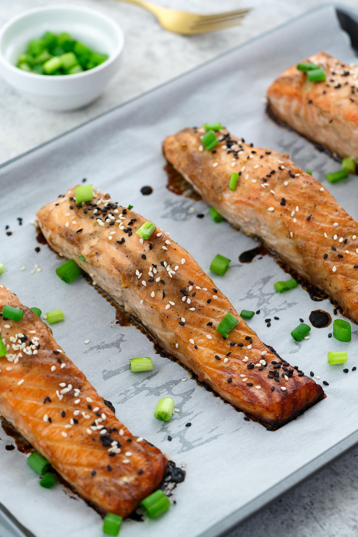 Miso-glazed salmon on a baking sheet garnished with black and white sesame seeds and spring onions. Surrounding the salmon are a golden fork and a cup of chopped spring onions.