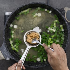 Dashi with green onions in a pot, dissolving miso paste using a fine mesh strainer.