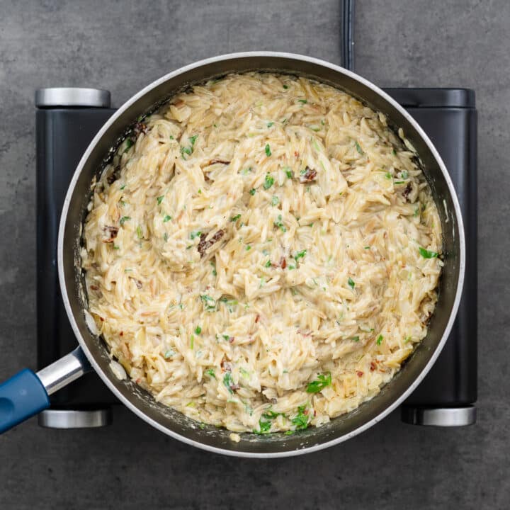 A wide-bottom pan containing creamy Orzo garnished with dried tomatoes, parmesan cheese, and parsley.