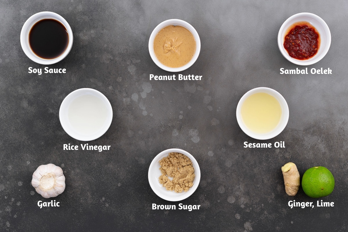 Ingredients for peanut sauce arranged on a gray table, including soy sauce, peanut butter, sambal oelek, rice vinegar, sesame oil, garlic, brown sugar, ginger, and lime.