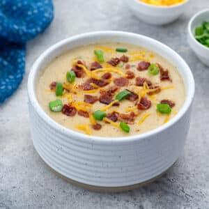 A bowl of creamy potato soup garnished with bacon and cheese, set on a white table. Nearby, a blue towel, a cup of extra cheese and chopped spring onions are arranged.