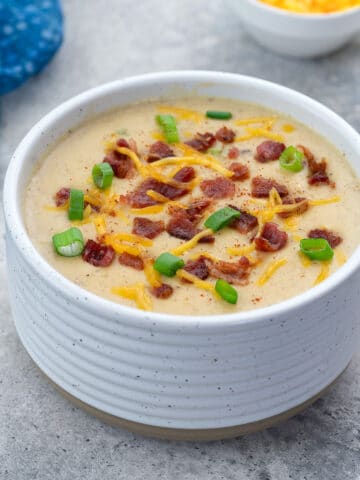 A bowl of creamy potato soup garnished with bacon and cheese, set on a white table. Nearby, a blue towel, a cup of extra cheese and chopped spring onions are arranged.