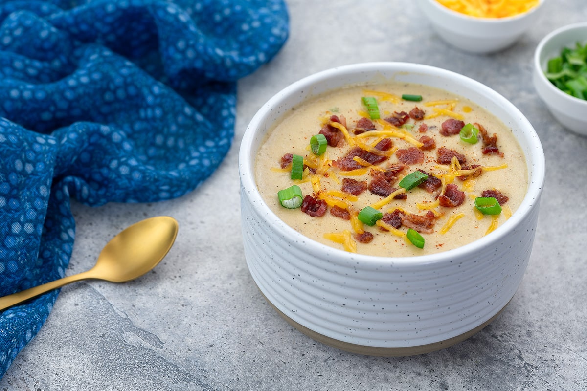 A bowl of creamy potato soup garnished with bacon and cheese, set on a white table. Nearby, a blue towel, a cup of extra cheese, chopped spring onions, and a golden spoon are arranged.