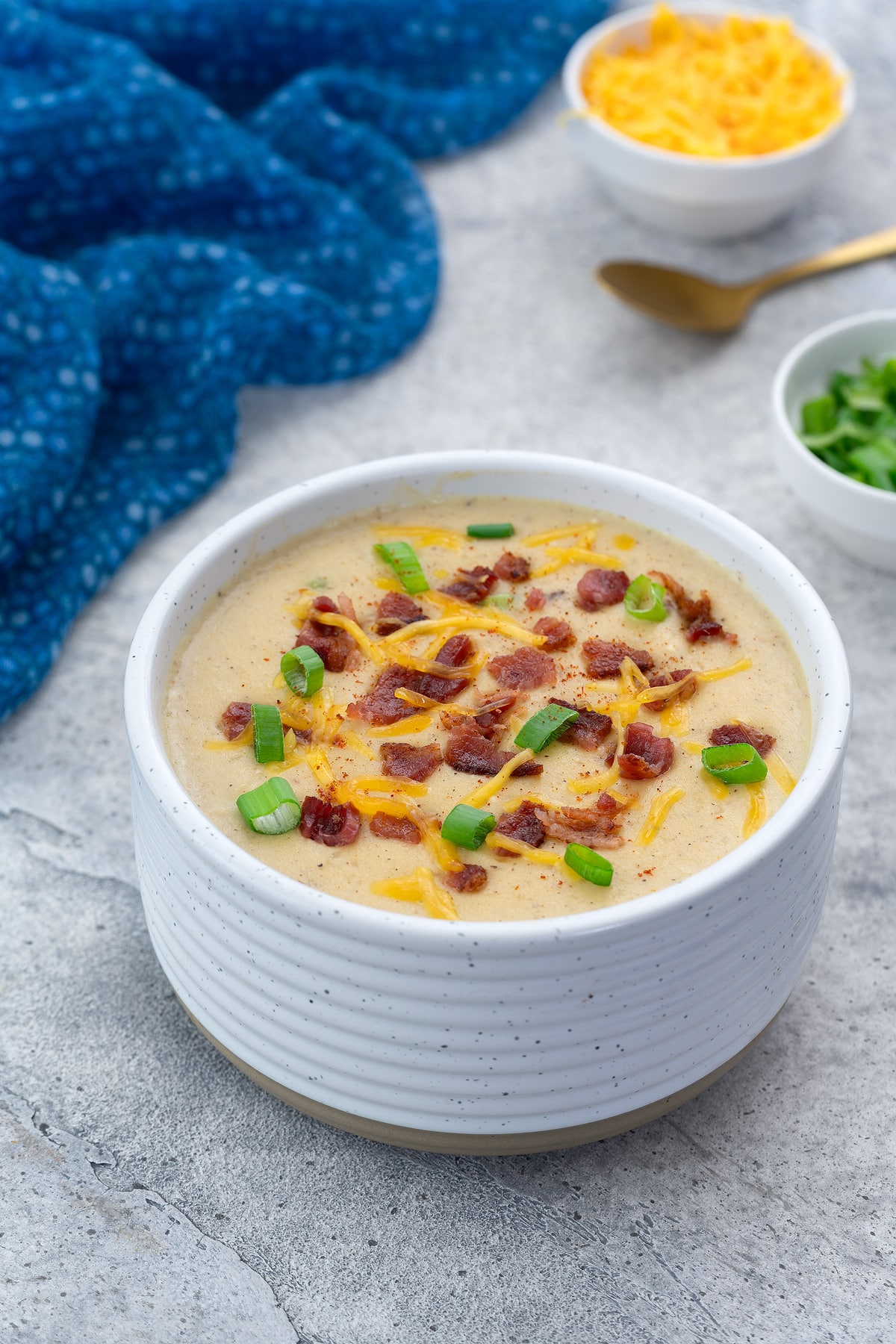 A bowl of creamy potato soup garnished with bacon and cheese, set on a white table. Nearby, a blue towel, a cup of extra cheese, chopped spring onions, and a golden spoon are arranged.