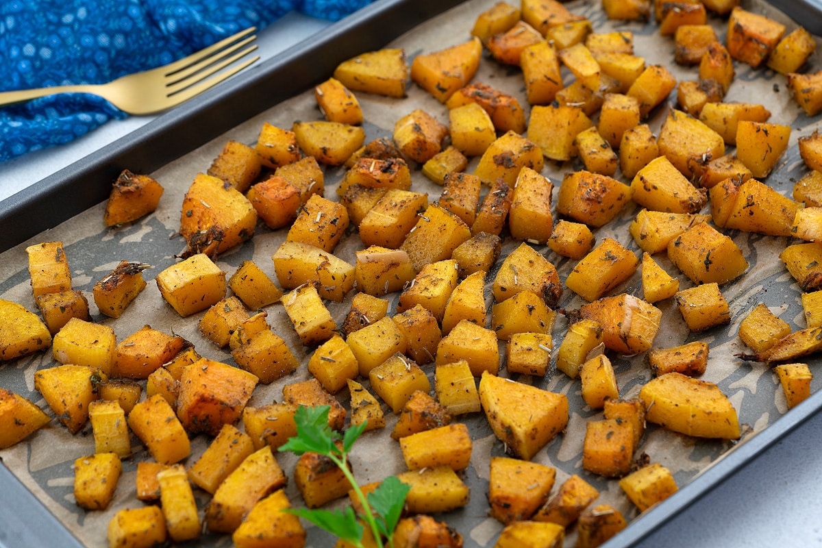 Roasted butternut squash in a baking tray, set on a white table. A fork and a blue towel are neatly placed next to the tray.