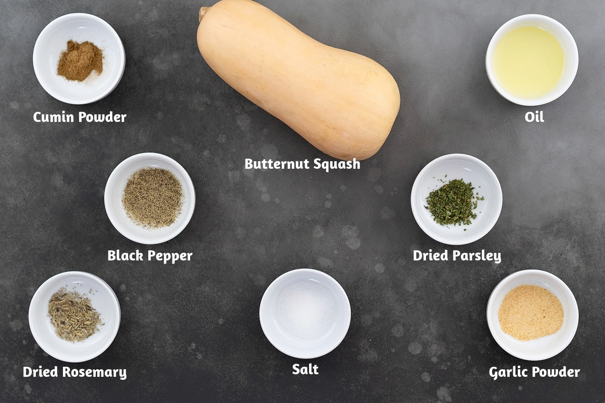 Ingredients for roasted butternut squash arranged on a gray table, including cumin powder, a whole butternut squash, oil, black pepper powder, dried parsley, dried rosemary, salt, and garlic powder.