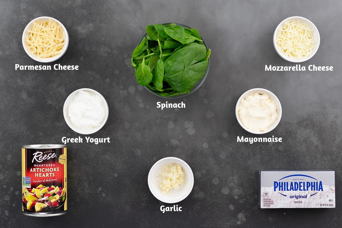 Ingredients for making spinach artichoke dip, parmesan cheese, spinach, mozzarella cheese, Greek yogurt, mayonnaise, artichokes, garlic, and cream cheese on a grey table.