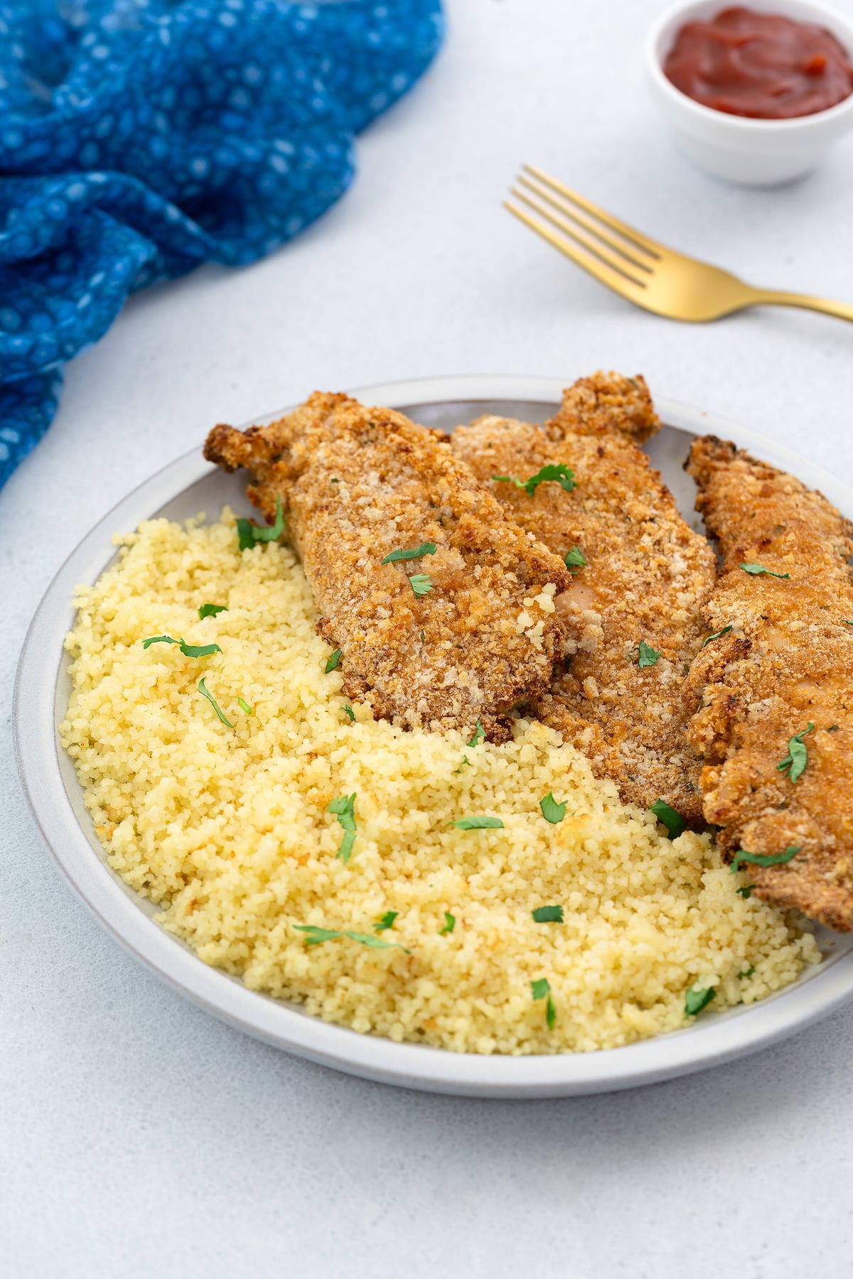 Air fryer chicken tenders on a white plate with couscous on a white table. A blue towel, a golden fork, and a cup of ketchup are placed around.