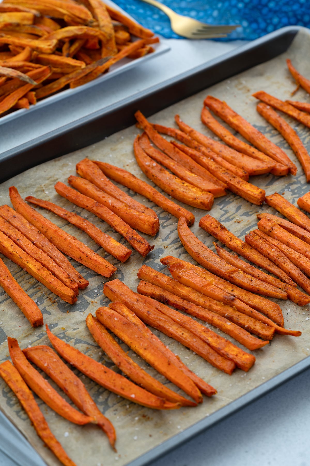 Baked sweet potato fries on a baking tray with a golden fork and a towel placed nearby.
