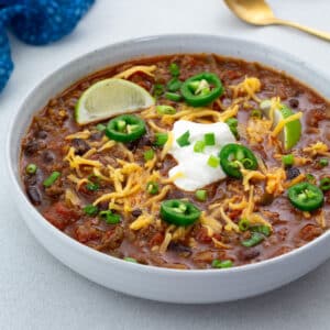 Beef chili in a white bowl on a white table with various toppings. A blue towel, and a golden spoon nearby.