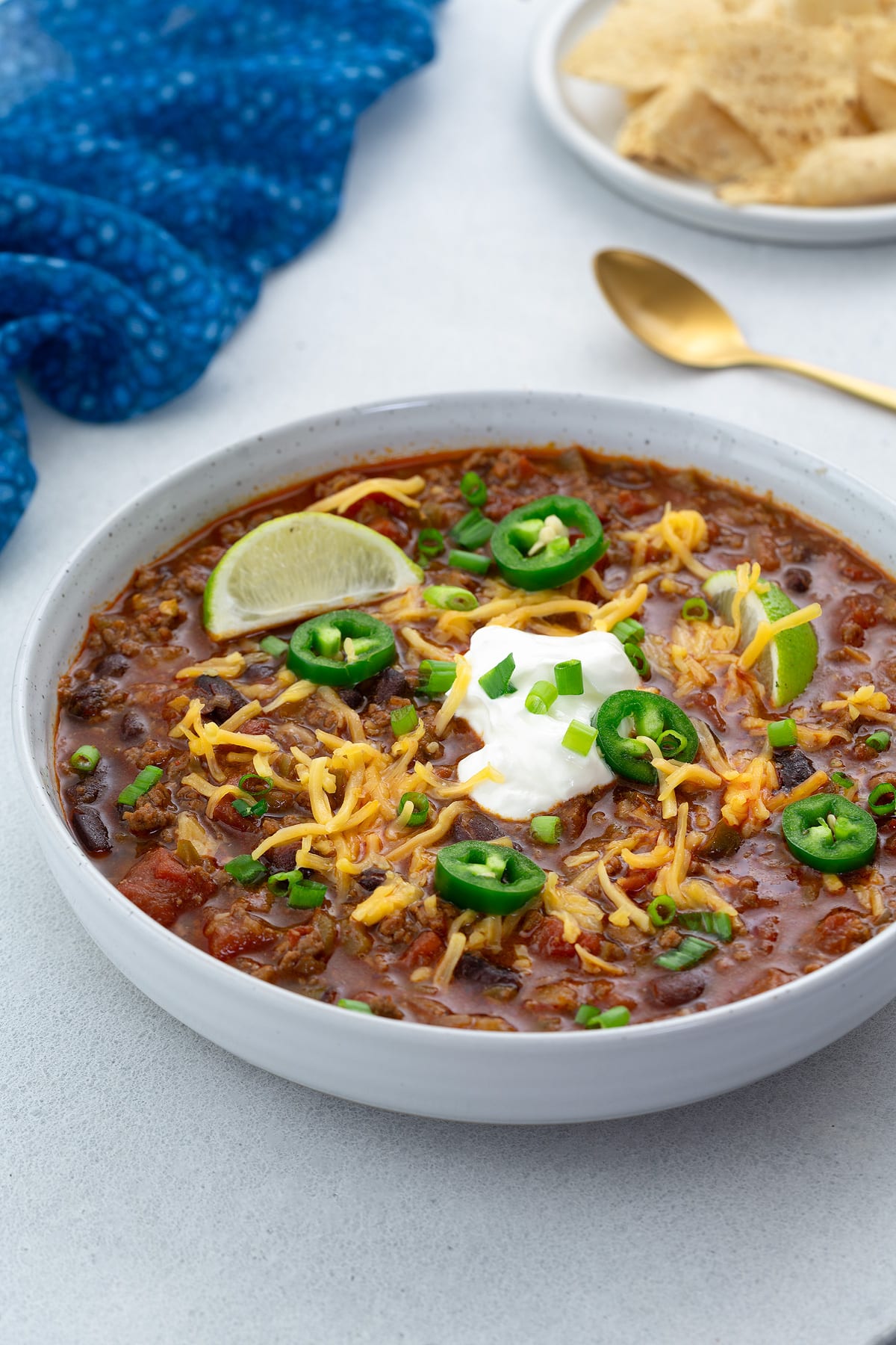 Beef chili in a white bowl on a white table with various toppings. A blue towel, chips on a plate, and a golden spoon nearby.