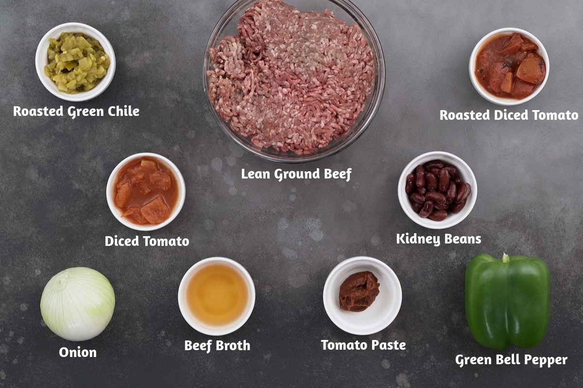 Ingredients for beef chili arranged on a gray table: roasted green chile, lean ground beef, roasted diced tomato, diced tomato, kidney beans, onion, beef broth, tomato paste, and green bell pepper.