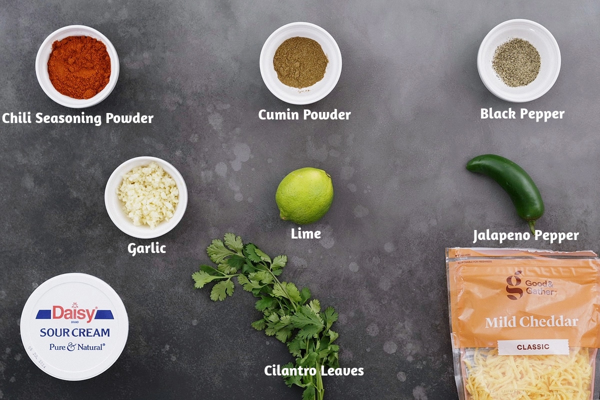 Ingredients for beef chili arranged on a gray table: chili seasoning powder, cumin powder, black pepper, garlic, lime, jalapeno pepper, sour cream, cilantro leaves, and cheddar cheese.