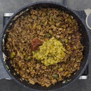 A pot with seasoned ground beef, tomato paste, and roasted green chilies.