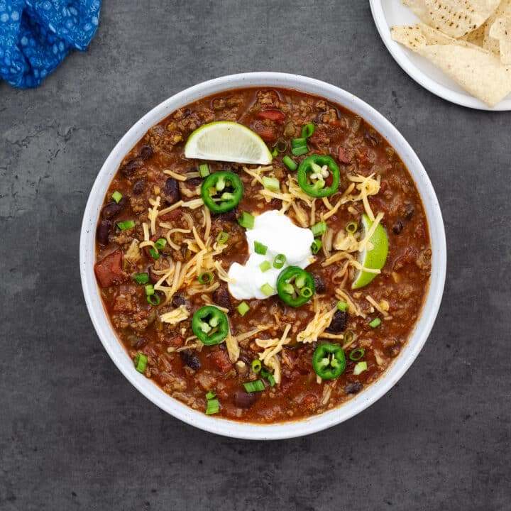 A bowl of beef chili topped with sour cream, cheddar cheese, lime wedges, cilantro leaves, and jalapenos.