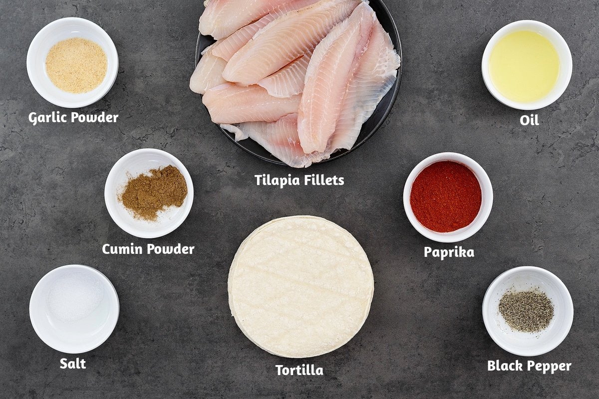 Ingredients for fish tacos placed on a gray table: garlic powder, tilapia fillets, oil, cumin powder, paprika, salt, tortillas, and black pepper.