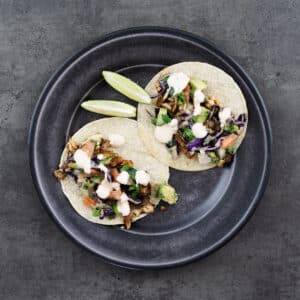 Plate with assembled tortillas featuring fried fish, vegetable topping, and taco sauce with lime wedges on the side.