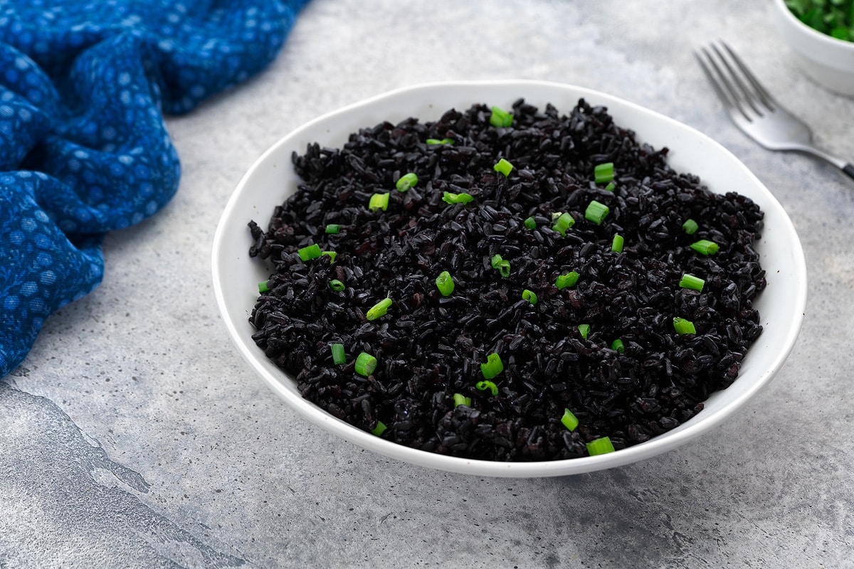 A white bowl of cooked forbidden black rice garnished with spring onion on a white table. Surrounding the bowl are a blue towel, a fork, and a cup of cut spring onion.