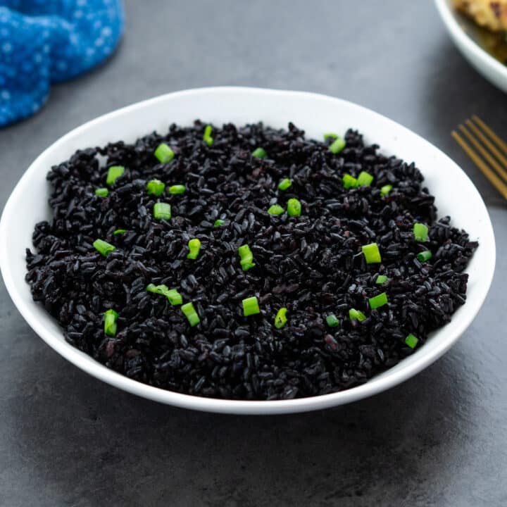 A bowl of cooked black rice garnished with spring onions, served alongside chicken.