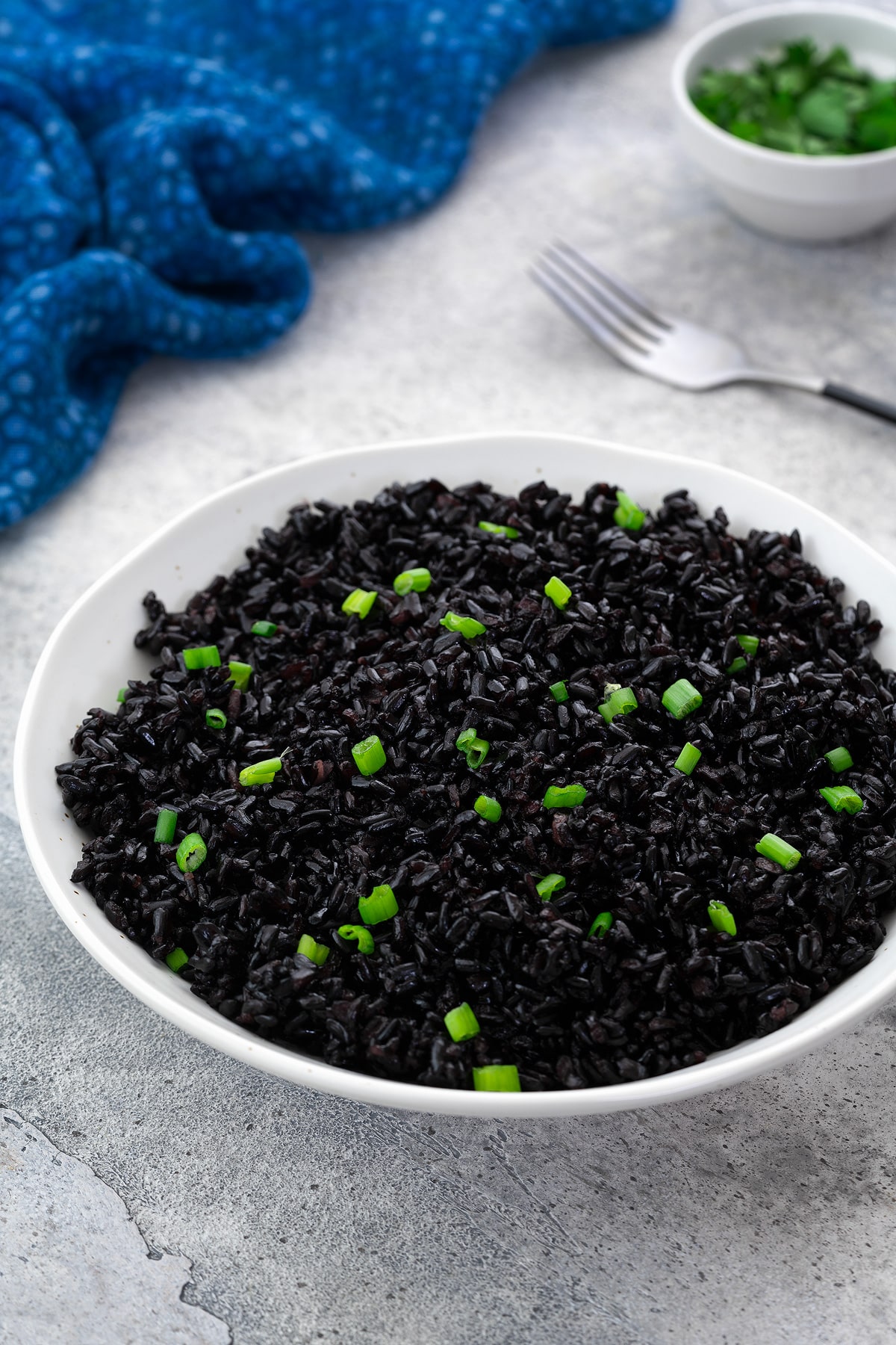 A white bowl of cooked forbidden black rice garnished with spring onion on a white table. Surrounding the bowl are a blue towel, a fork, and a cup of cut spring onion.