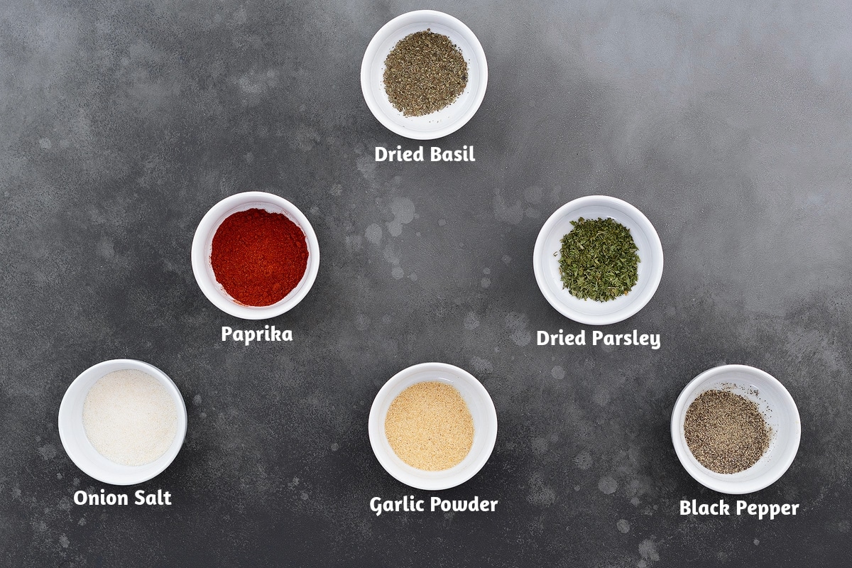 Ingredients for French fry seasoning arranged on a gray table, including dried basil, paprika, dried parsley, onion salt, garlic powder, and black pepper.