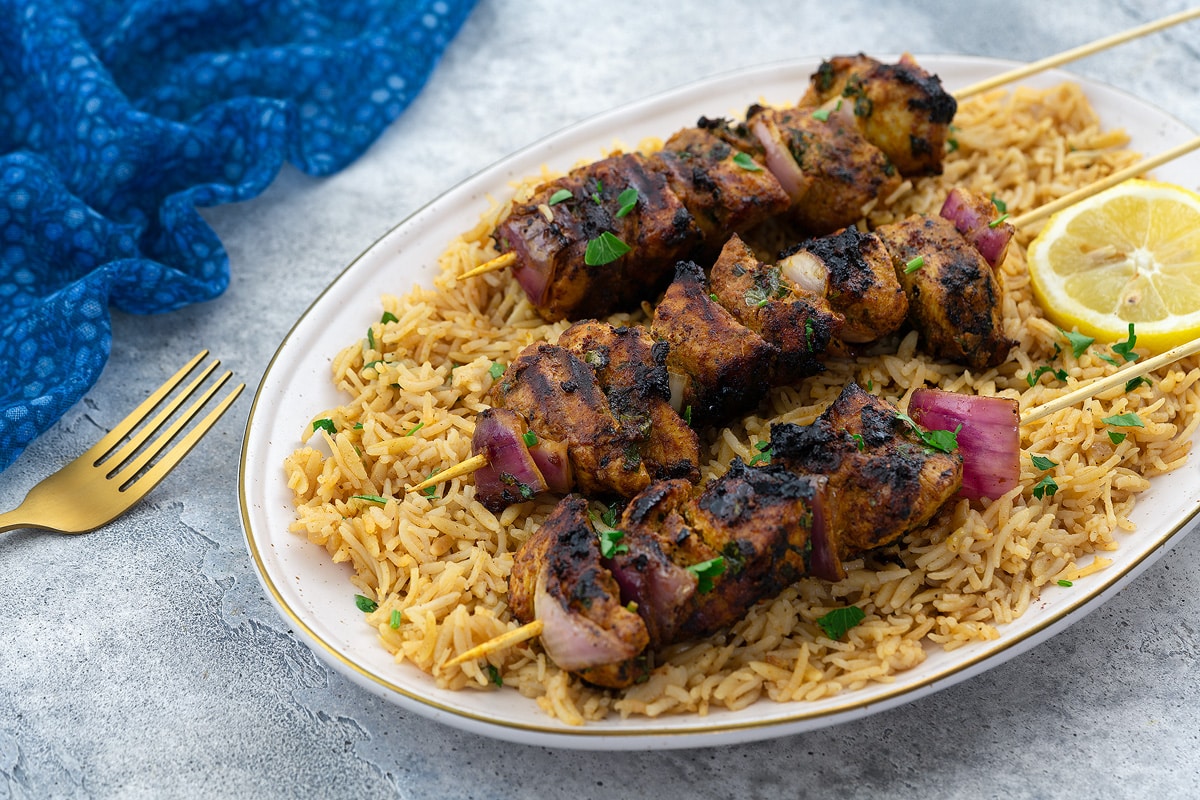 Grilled chicken kabobs on skewers served over a bed of fluffy rice pilaf, set on a white table. A blue towel and a golden fork are placed nearby.