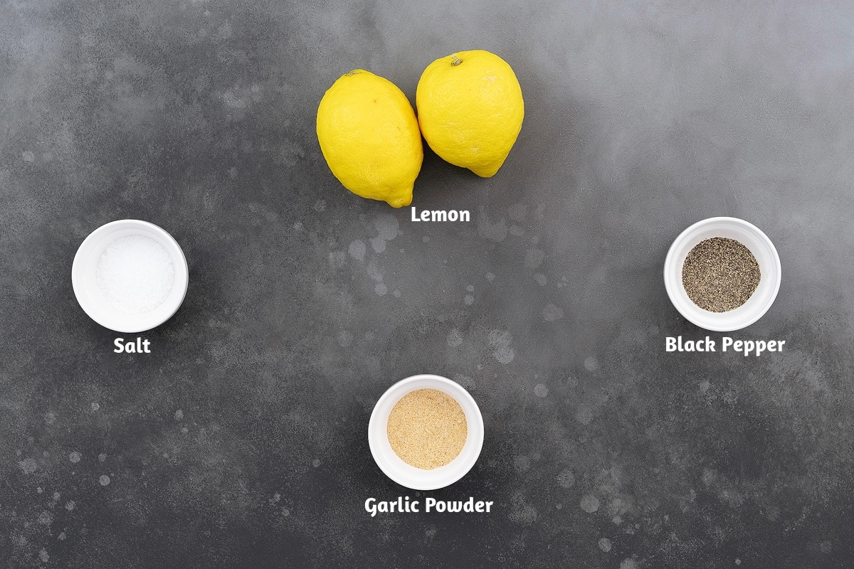 On a gray table, ingredients for lemon pepper seasoning are laid out: salt, lemon, black pepper, and garlic powder.