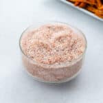 A glass bowl of seasoned salt on a white table, accompanied by a plate of sweet potato fries nearby.