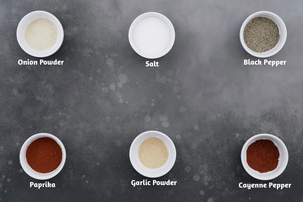 An array of seasoned salt ingredients displayed on a gray table: onion powder, salt, black pepper, paprika, garlic powder, and cayenne pepper.
