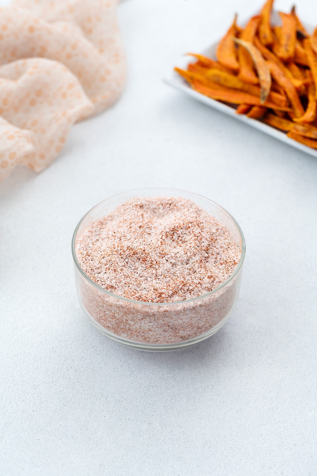 A glass bowl of seasoned salt on a white table, accompanied by a towel and a plate of sweet potato fries nearby.