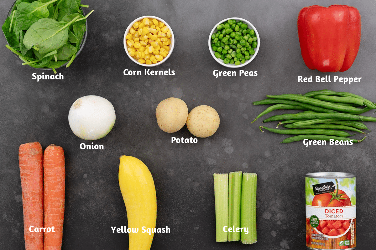 Ingredients for vegetable soup displayed on a gray table: spinach, corn kernels, green peas, red bell pepper, onion, potato, green beans, carrot, yellow squash, celery, and diced tomato.