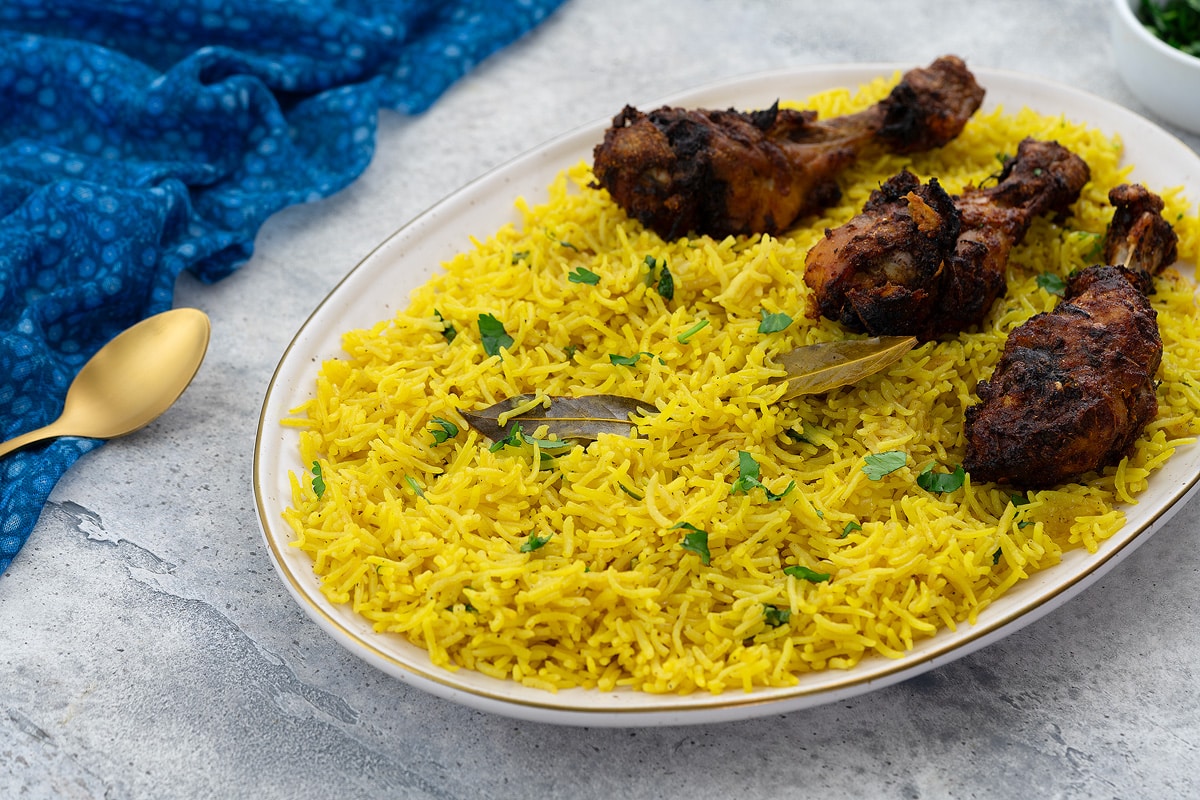 Yellow rice on an oval-shaped plate with chicken drumsticks. A blue towel, golden fork, and a cup of chopped cilantro leaves placed around.