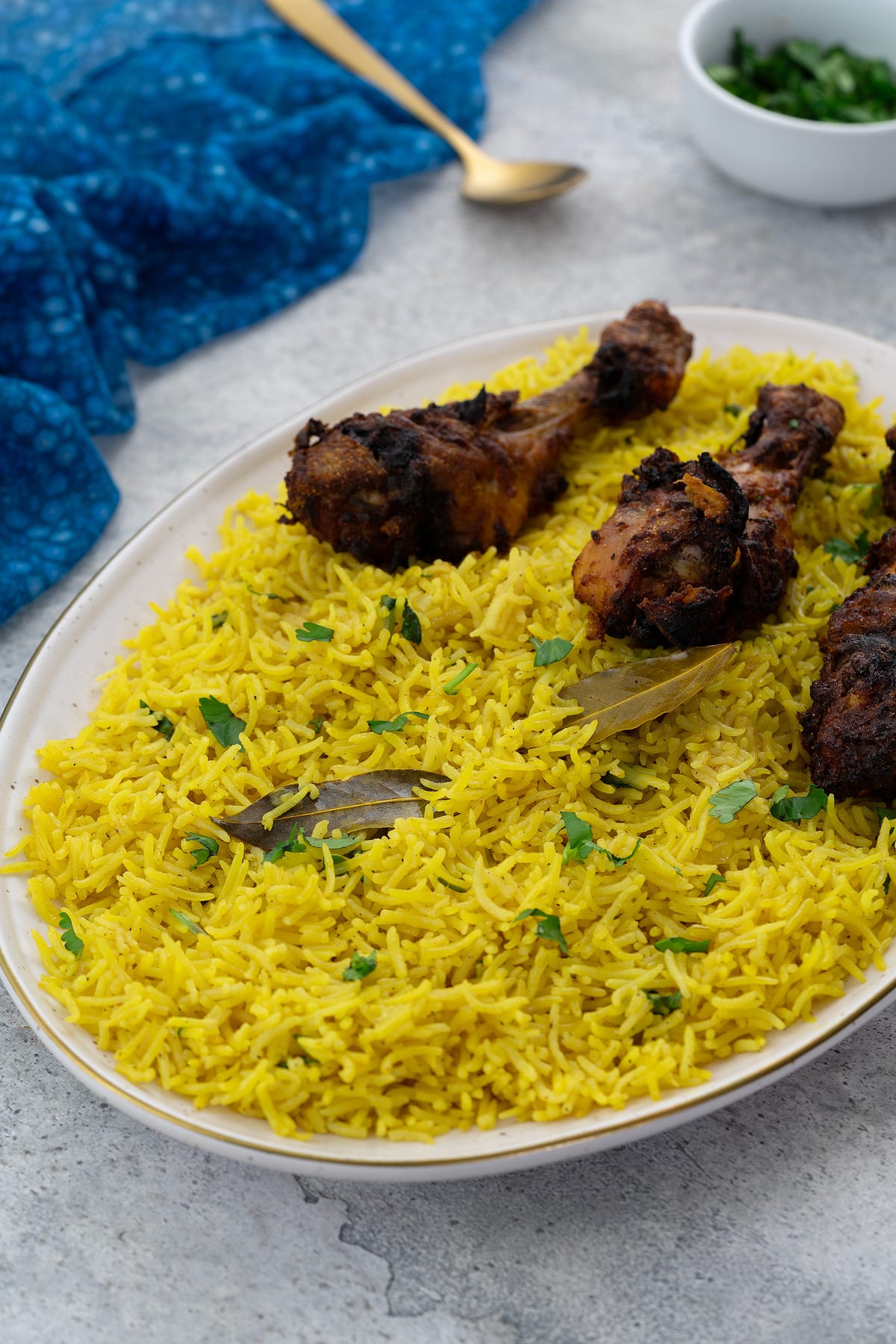 Yellow rice on an oval-shaped plate with chicken drumsticks. A blue towel, golden fork, and a cup of chopped cilantro leaves placed around.