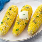 Boiled corn on the cob on an oval plate on a white table, with a blue towel placed nearby.
