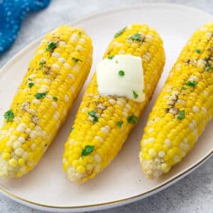 Boiled corn on the cob on an oval plate on a white table, with a blue towel placed nearby.