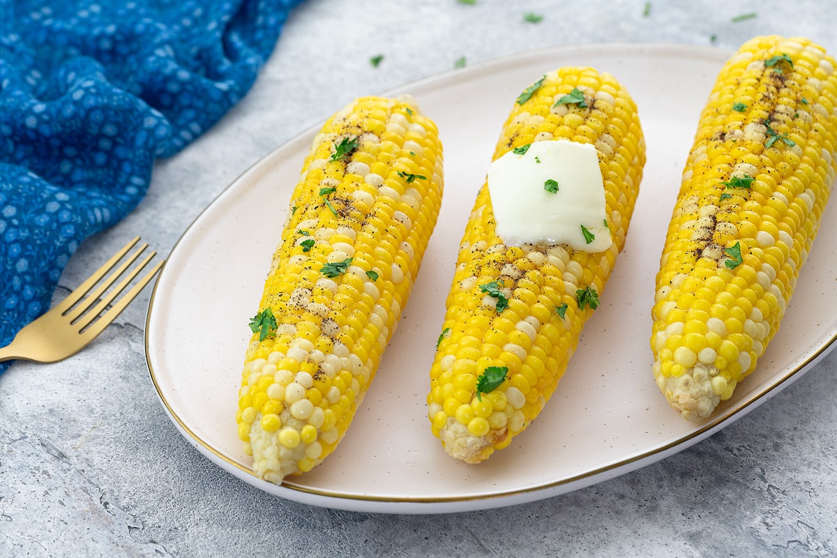 Boiled corn on the cob on an oval plate on a white table. Surrounding items include a blue towel, a golden fork, and a cup of cilantro.