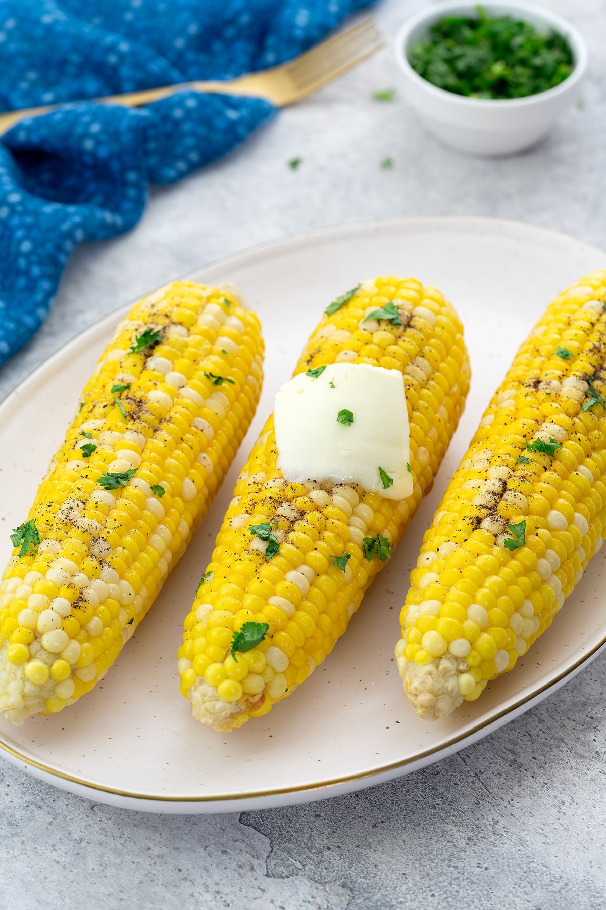 Boiled corn on the cob on an oval plate on a white table. Surrounding items include a blue towel, a golden fork, and a cup of cilantro.