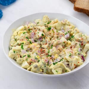 Creamy egg salad in a white bowl on a white table, with a blue towel and slices of bread neatly arranged at the top corners.