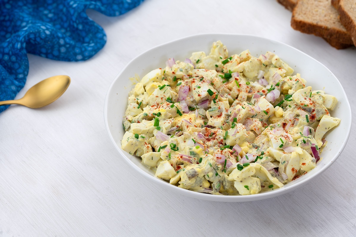 Creamy egg salad served in a white bowl on a white table, accompanied by a blue towel, a golden spoon, and slices of bread arranged around the bowl.