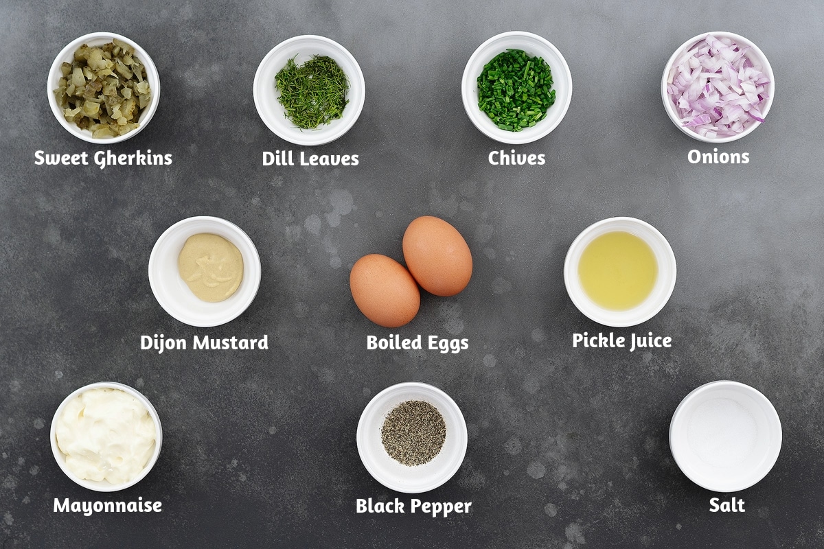 Ingredients for egg salad recipe displayed on a gray table: sweet gherkins, dill leaves, chives, onions, Dijon mustard, boiled eggs, pickle juice, mayonnaise, black pepper, and salt arranged in groups.