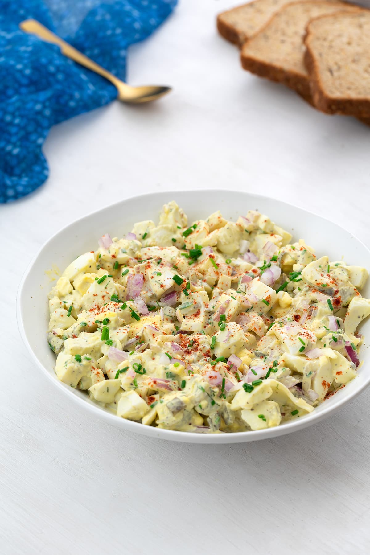 Creamy egg salad served in a white bowl on a white table, accompanied by a blue towel, a golden spoon, and slices of bread arranged around the bowl.