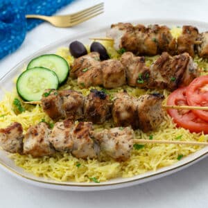 Grilled Greek Chicken Souvlaki skewers served on a bed of yellow rice, arranged on an oval plate on a white table. A blue towel and a golden fork are placed nearby.