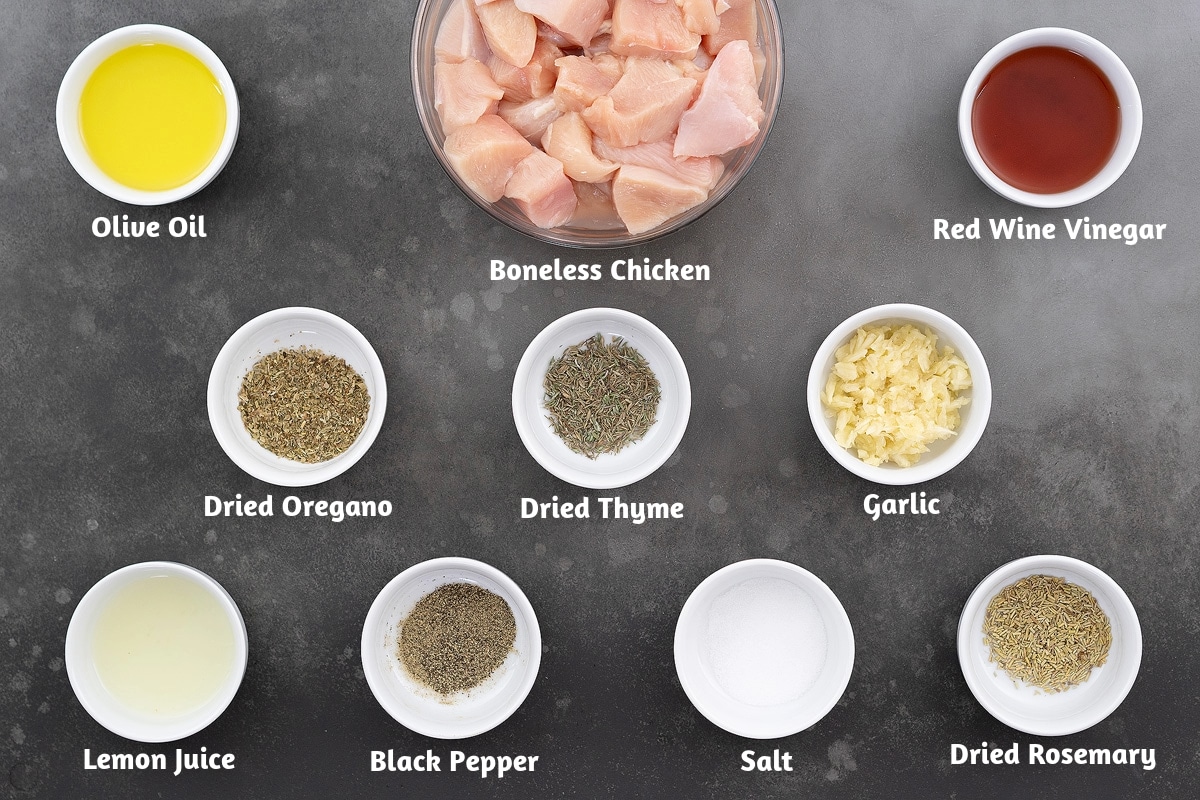 Ingredients for Greek Chicken Souvlaki on a gray table, including olive oil, boneless chicken, red wine vinegar, herbs, garlic, lemon juice, and spices.