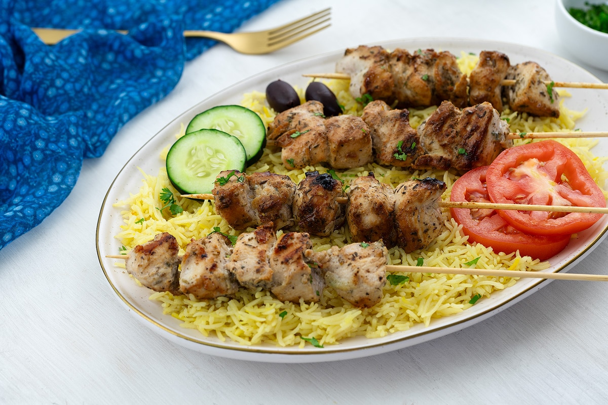 Grilled Greek Chicken Souvlaki skewers served on a bed of yellow rice, arranged on an oval plate on a white table. A blue towel, a golden fork, and a cup of fresh cucumber and tomato slices are placed nearby.