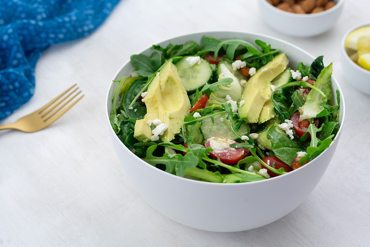 Green salad in a white bowl on a white table, surrounded by a cup of almonds, lemon slices, a blue towel, and a golden fork.